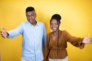 Genç Afrikalı Amerikalı çift sarı arka planda durup kameraya bakıp kucaklaşmak için kollarını açıp gülümsüyor.