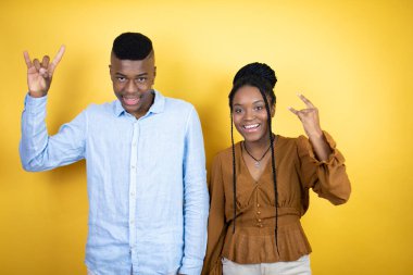 Genç Afro-Amerikan çifti sarı arka planda durup çılgın bir ifadeyle el kaldırarak rock sembolü yapıyor.
