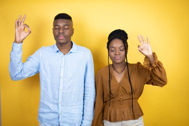 Genç Afro-Amerikan çifti sarı arka planda dikiliyor rahatlıyor ve gözleri kapalı gülümsüyor parmaklarıyla meditasyon yapıyor.