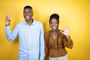 Genç Afro-Amerikan çifti sarı arka planda duruyor ve yedinci parmaklarını gösterip gülümserken mutlu ve kendinden emin