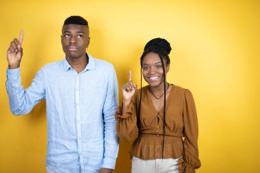 Genç Afro-Amerikan çift sarı arka planda durup gülümsüyor, kameraya bakıyor ve parmaklarıyla işaret ediyor ve kollarını kaldırıyor.
