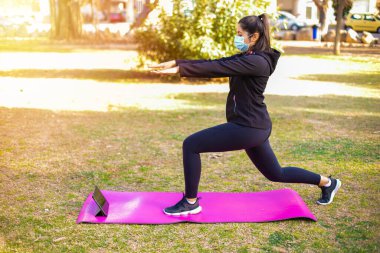 Maskeli genç esmer sporcu kadın parkta paspas üzerinde egzersiz yapıyor ve tabletteki spor videosuna bakıyor.