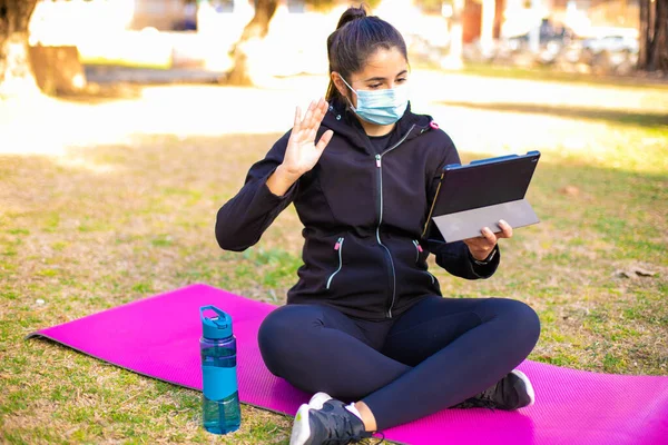 Young brunette sportswoman with mask doing exercise on a mat in the park using tablet and saying hello