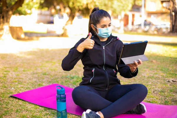 Young brunette sportswoman with mask doing exercise on a mat in the park using tablet and doing ok sign, thumb up with fingers, excellent sign