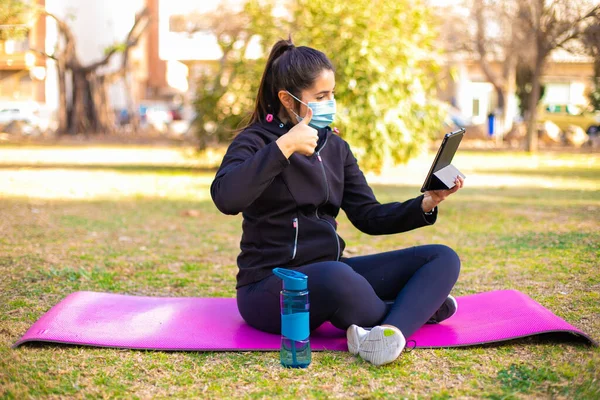 Young brunette sportswoman with mask doing exercise on a mat in the park using tablet and doing ok sign, thumb up with fingers, excellent sign