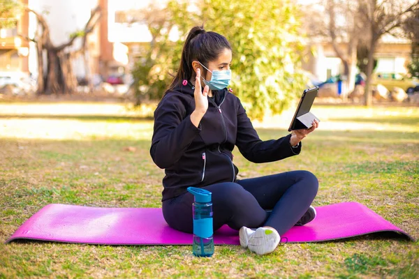 Young brunette sportswoman with mask doing exercise on a mat in the park using tablet and saying hello