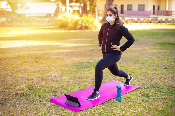 Young brunette sportswoman with mask doing exercise on a mat in the park looking at sport video on tablet