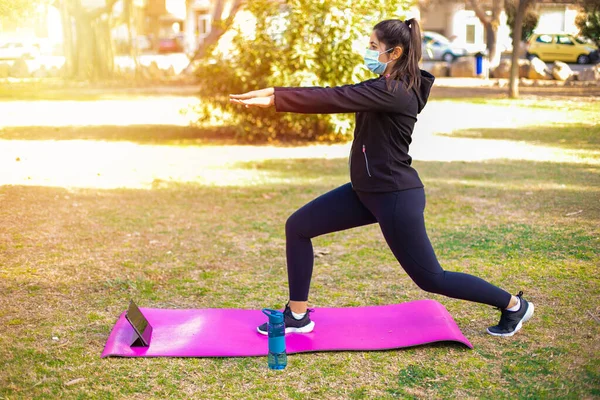 Young brunette sportswoman with mask doing exercise on a mat in the park looking at sport video on tablet