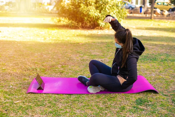 Young brunette sportswoman with mask doing exercise on a mat in the park looking at sport video on tablet