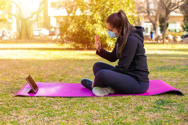 Young brunette sportswoman with mask doing exercise on a mat in the park using tablet and saying hello