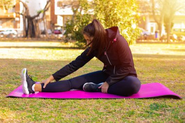 Genç esmer sporcu kadın parkta paspasın üstünde egzersiz yapıyor.