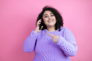 Genç, güzel bir kadın, pembe arka plan üzerine günlük süveter giyip akıllı telefondan konuşurken mutlu bir gülümsemeyle el ve parmağı işaret ediyor.