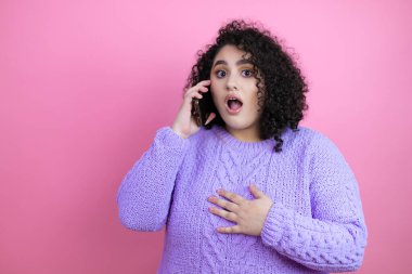 İzole edilmiş pembe arka plan üzerine sıradan süveter giyen genç ve güzel bir kadın telefonda endişeli bir ifadeyle konuşuyor.