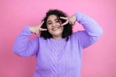 İzole edilmiş pembe arka plan üzerine günlük süveter giyen genç ve güzel bir kadın. Yüzünde parmakları olan barış sembolü yapıyor. Gülümsüyor, zaferini gösteriyor.