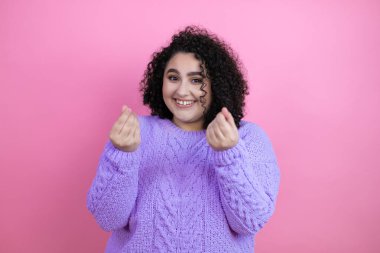 İzole edilmiş pembe arka plan üzerine günlük süveter giyen genç güzel bir kadın. Elleriyle para hareketi yapıyor. Maaş ödemesi istiyor.