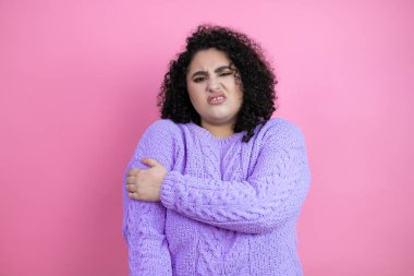 Genç, güzel bir kadın, pembe arka plan üzerine günlük süveter giyiyor. Omzunda ağrı ve acı dolu bir ifade var.