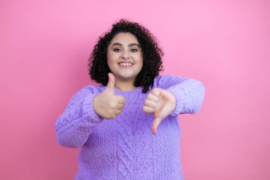 Genç ve güzel bir kadın pembe arka plan üzerine günlük süveter giyip baş parmak kaldırıyor, anlaşmazlık ve mutabakat ifadesi takınıyor. Çılgın çatışma