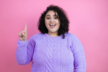 İzole edilmiş pembe arka plan üzerine sıradan süveter giyen genç ve güzel bir kadın parmağını kafasında gezdirmiş ve bir fikri olduğunu düşünüyor..