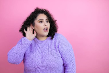 İzole edilmiş pembe arka plan üzerine günlük süveter giyen genç ve güzel bir kadın dedikodu ya da dedikodulara kulak misafiri oldu.