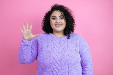 İzole edilmiş pembe arka plan üzerine sıradan süveter giyen genç ve güzel bir kadın beş numaralı parmaklarıyla işaret ederken kendinden emin ve mutlu gülümsüyor.