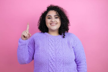 İzole edilmiş pembe arka plan üzerine sıradan süveter giyen genç ve güzel bir kadın kendini beğenmiş ve mutlu bir şekilde gülümserken bir numaralı parmaklarıyla işaret ediyor.