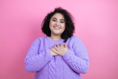İzole edilmiş pembe arka planda rahat bir süveter giyen genç ve güzel bir kadın. Elleri göğsünde ve yüzünde minnet dolu bir jest ile gülümsüyor..
