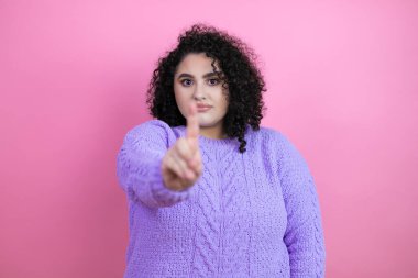 İzole edilmiş pembe arka plan üzerine sıradan süveter giyen genç ve güzel bir kadın bir numaralı parmaklarıyla işaret ediyor.