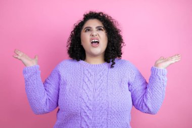 İzole edilmiş pembe arka plan üzerine sıradan bir süveter giyen genç, güzel bir kadın. Kolları ve elleri havada, kafası karışmış bir ifade.