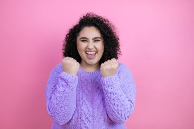 Genç, güzel bir kadın pembe arka plan üzerine günlük süveter giyiyor. Çok mutlu ve heyecanlı. Kollarını kaldırmış, gülümseyen ve başarı için çığlık atan kazanan bir jest yapıyor..