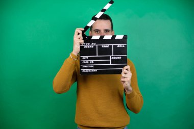 Yeşil arka plan üzerinde günlük kıyafetler giyen yakışıklı bir adam elinde klaket tutarak çok mutlu.