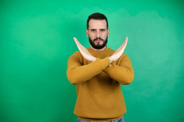 Yeşil arka planda rahat kıyafetler giyen yakışıklı bir adam. Reddedilme ifadesi. Kollarını kavuşturma, negatif işaret, kızgın surat.