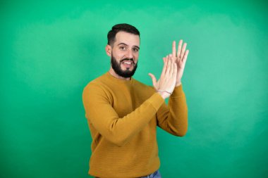 Yeşil arka planda rahat kıyafetler giyen yakışıklı adam alkışlıyor ve mutlu ve mutlu, gururlu elleri bir arada alkışlıyor.