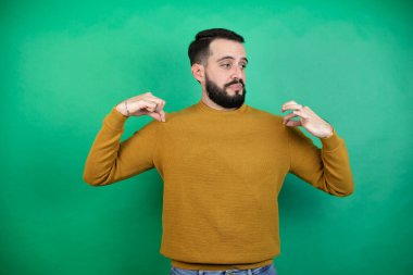 Yeşil arka plan üzerinde günlük kıyafetler giyen yakışıklı bir adam tişörtünü tutarken başarılı bir yüz ifadesi takınıyor.