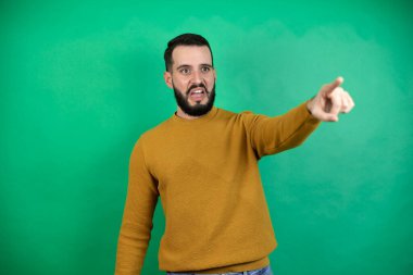 Yeşil arka plan üzerinde sıradan kıyafetler giyen yakışıklı bir adam. Parmağını ileri doğru uzatmış, ağzı açık, şaşırmış, ön tarafta bir şey var.