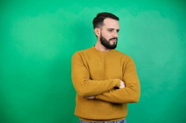 Yeşil arka planda rahat kıyafetler giyen yakışıklı adam yorgun ve kollarını kavuşturmaktan sıkılmış görünüyor.