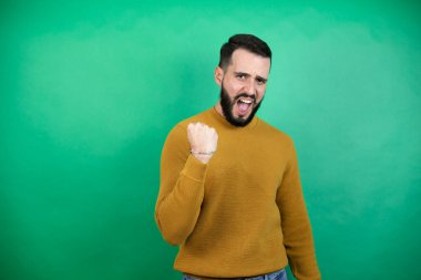 Yeşil arka planda rahat kıyafetler giyen yakışıklı bir adam öfkeyle bağırırken kızgın ve kızgın bir şekilde yumruk kaldırıyor. Öfke ve agresif kavram.