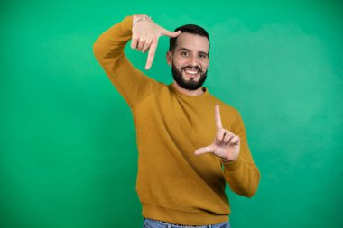 Yeşil arka planda rahat kıyafetler giyen yakışıklı bir adam gülümseyerek elleri ve parmakları mutlu yüzlü bir çerçeve yapıyor.