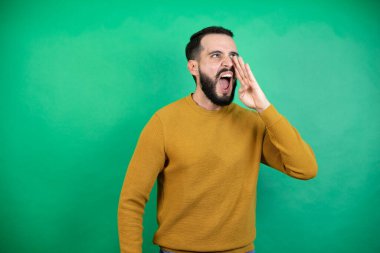 Yeşil arka planda rahat kıyafetler giyen yakışıklı adam elleri ağızda bağırarak ve yüksek sesle bağırıyor.