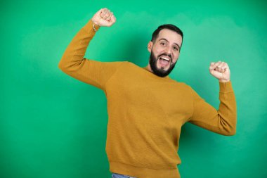 Yeşil arka planda rahat kıyafetler giyen yakışıklı bir adam mutlu ve heyecanlı. Kollarını kaldırıp, gülümseyip başarı için bağırarak kazanan bir jest yapıyor..