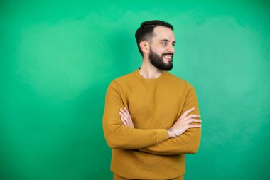 Yeşil arka planda rahat kıyafetler giyen yakışıklı bir adam kollarını kavuşturmuş, ikna olmuş ve kendinden emin bir şekilde yana bakıyor.