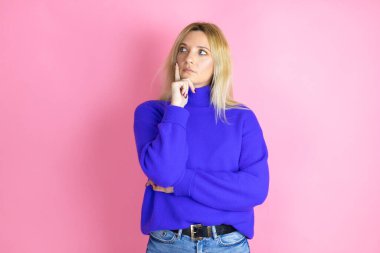 İzole edilmiş pembe arka plan üzerine günlük süveter giyen genç ve güzel bir kadın. Ciddi bir yüz ifadesi. Çene üstüne el ile soru sormayı düşünen, kafa karıştırıcı bir fikir.