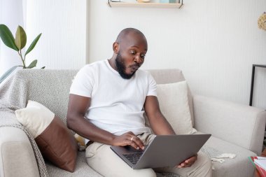 Beyaz tişörtlü afro-amerikan bir adam boş zamanlarını oturma odasında kanepede oturup dizüstü bilgisayarıyla film izleyerek geçiriyor. İnsanlar, dinlenme ve teknoloji kavramı.