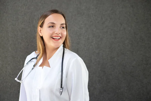 医療コートの魅力的な若い女性医師の横のクローズアップ写真は 遠くを見て 広く笑みを浮かべて 灰色の背景に隔離されたスペースをコピーします 人々の概念 — ストック写真