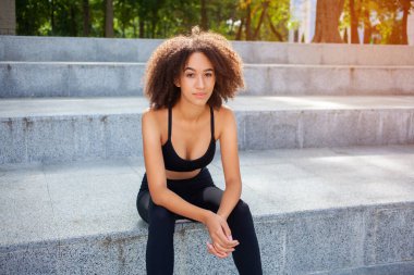 Siyah spor tişörtlü, kıvırcık tenli genç bir kızın üst köşesinde parlayan bir fotoğraf ve merdivenlerde tozluklar, şehir parkında yarıştan sonra dinleniyor. Spor, aktif dinlenme, sağlıklı konsept.