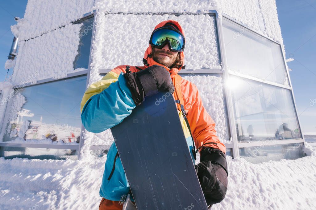 Happy Male snowboarder wearing coloured clothing standing near snow covered building on sunny day. Holidays on ski resort