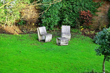 Çimlerin üzerinde iki beton sandalye ve masa, çalılarla çevrili, çit, tuğla.