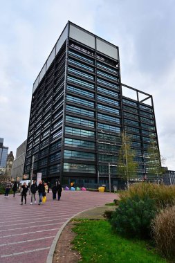 Yansıtıcı şebeke cephesi, yaya meydanı, heykeller, yeşillik ve saat kulesi görünür..