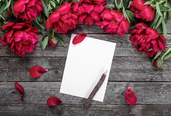 Vitt papper kort med penna på rustik trä bakgrund med rosa pioner och kronblad. Blommor. Arbetsplats. Blank för hälsningar — Stockfoto