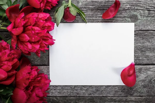 Cartão de papel branco sobre fundo de madeira rústica com peônias e pétalas rosa. Flores. Espaço de trabalho. Espaço em branco para saudações — Fotografia de Stock