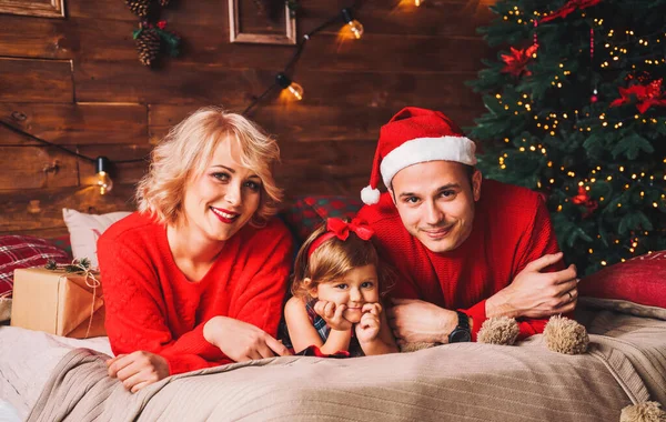 戴着圣诞圣诞礼帽的家人躺在床上 父亲和婴儿在由圣诞树装饰的客厅里玩乐 并赠送礼品盒 新年及圣诞节假期 — 图库照片